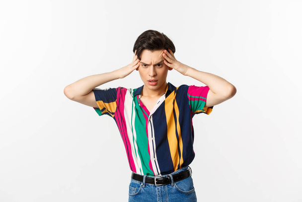 Anxious young gay man standing in panic, holding hands on head and staring down troubled, having a problem, standing over white background - Фото, изображение