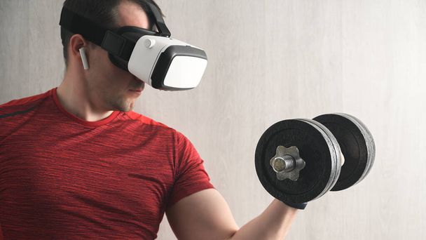 Fitness, sport and technology. Portrait of concentration young adult sport man in a red t-shirt training in home, holding dumbbell with arm, doing exercises for biceps and looking at vr glasses - Φωτογραφία, εικόνα