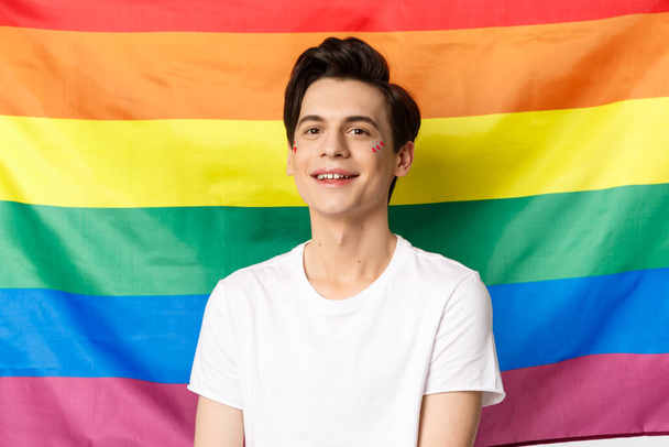 Krásný gay muž s třpytkami na tváři, usmívá šťastný a hrdý na kameru, stojí proti duhové vlajce hrdosti, komunitní práva lgbtq a lidé koncept - Fotografie, Obrázek