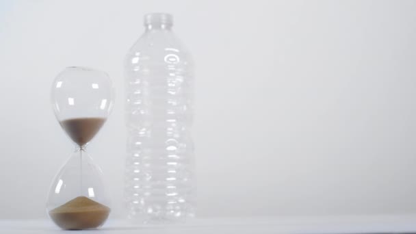 Ένα ευρύ πλάνο ενός μισογεμάτου χρονοδιακόπτη άμμου δίπλα σε ένα πλαστικό μπουκάλι μίας χρήσης σε λευκό φόντο - Πλάνα, βίντεο