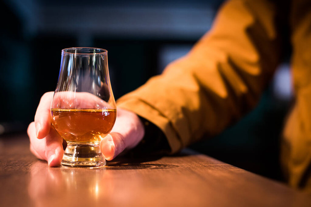 Kleur close-up schot een hand met een Glencairn whisky glas op een houten tafel, met ondiepe scherptediepte. - Foto, afbeelding