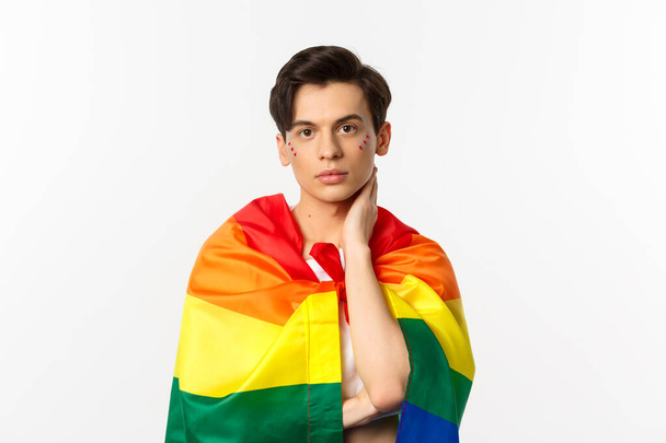 Waist-up shot of beautiful gay man with glitter on face, wrap himself with rainbow lgbt flag and touching face gently, looking at camera, white background - Фото, изображение