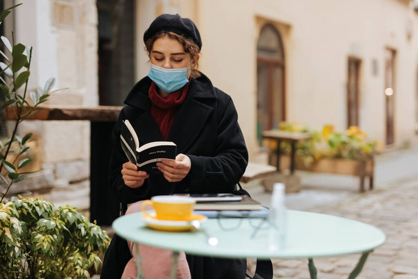 Stunning young woman in medical hygiene mask sitting at cafe table and reading book outdoors. Prevention of coronavirus outbreak. - Photo, Image