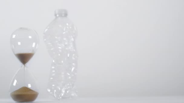 白地に砕いた単用途プラスチックボトルの横に半分の砂の砂時計の広いショット - 映像、動画