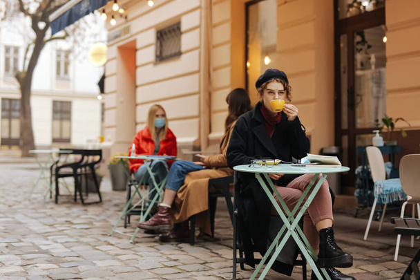 Стильная молодая леди сидит одна на открытой террасе кафе и пьет свежий кофе. Фон двух женщин, сидящих за столом в медицинских масках. Профилактика коронавируса. - Фото, изображение