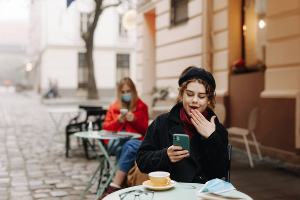 Довольно молодая брюнетка в стильном теплом наряде сидит на террасе кафе на открытом воздухе и улыбается, глядя на экран смартфона. Лежащая на столе медицинская маска и дезинфицирующее средство. - Фото, изображение