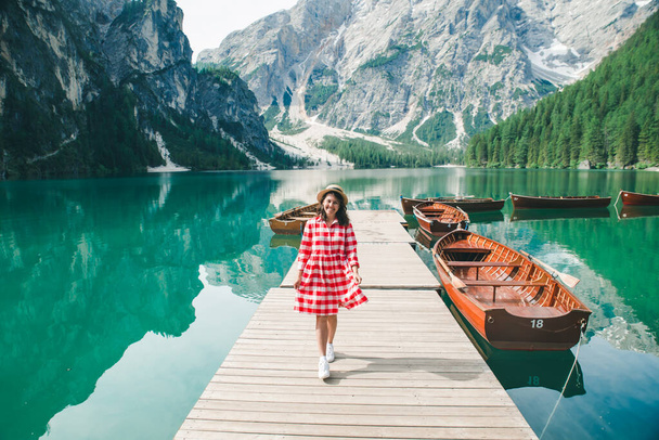 Молодая красивая женщина красный сарафан прогуливаясь по деревянной пристани с лодками. летние каникулы. горное озеро на заднем плане - Фото, изображение