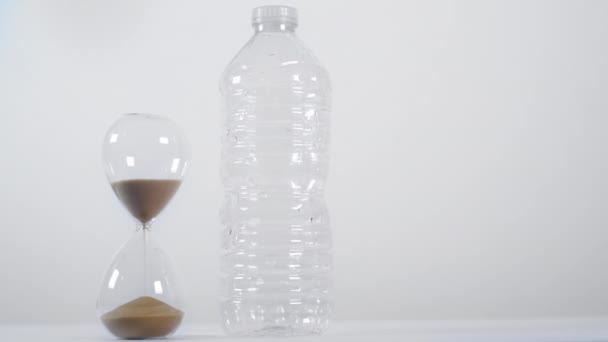 Plan large d'une minuterie sable à moitié pleine à côté d'une bouteille en plastique à usage unique sur fond blanc  - Séquence, vidéo