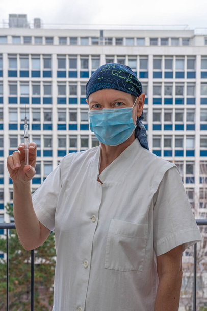 Paris, Frankreich - 31 01 2021: Maskierte Krankenschwester in weißem Laborkittel mit blauem Kopftuch und Spritze in der Hand - Foto, Bild