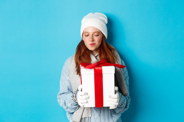Kış tatili ve Noel satış konsepti. İlgi çekici kızıl saçlı kız hediyelerle dolu kutuya bakıyor, tahmin et içinde ne var, mavi arka planda duruyor. - Fotoğraf, Görsel