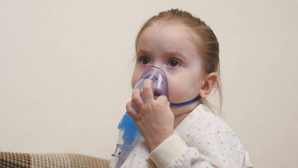 マスクの可愛い赤ん坊が吸入器を通して呼吸している。ホーム吸入手順.子供はネブライザーで呼吸療法を受けています。自宅でコロナウイルスのために赤ちゃんを治療してください。小さな子供 - 写真・画像