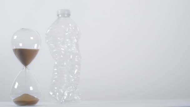 Un ampio scatto di un timer a sabbia piena accanto a una bottiglia di plastica monouso su uno sfondo bianco - Filmati, video