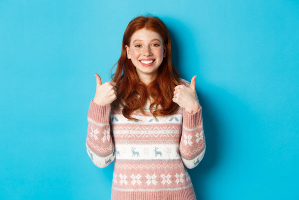 Chica pelirroja alegre en suéter de invierno mostrando los pulgares hacia arriba, de acuerdo y como, mostrando su apoyo y sonriendo, de pie satisfecho contra el fondo azul - Foto, imagen