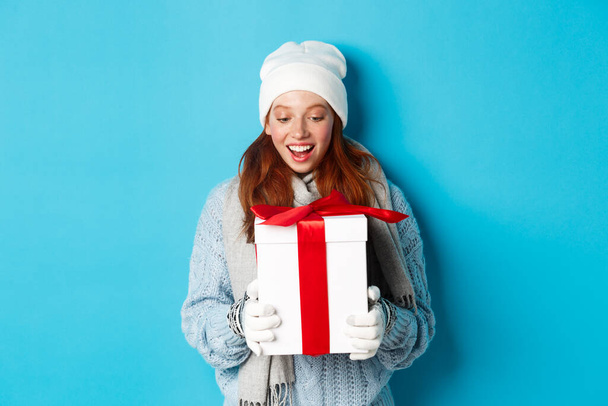 Winter holidays and Christmas eve concept. Surprised cute redhead girl in beanie and sweater receiving New Year gift, looking at present amazed, standing over blue background - Foto, immagini