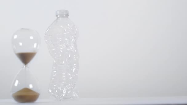 Um tiro largo de um temporizador de areia meio cheio ao lado de uma garrafa de plástico de uso único esmagada em um fundo branco - Filmagem, Vídeo