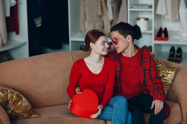 Sevgililer günü hediyesi olan gülümseyen genç kadın ve erkek. Ev kanepede kırmızı kalp şeklinde. - Fotoğraf, Görsel