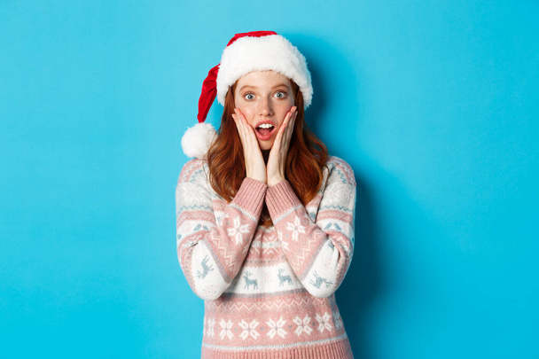 Winter and Christmas Eve concept. Impressed redhead girl in santa hat hear amazing news, touching cheeks and open mouth fascinated, standing against blue background - Photo, Image