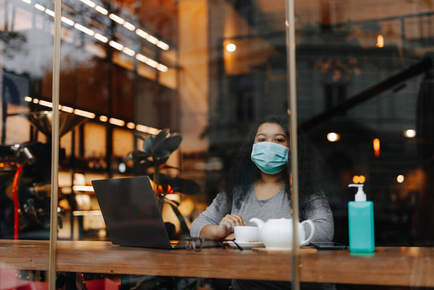 居心地の良いカフェに座っている間、アフリカの女性のフリーランサーはノートパソコンで好きな仕事を楽しんでいます。公共の場所で医療保護マスクを身に着けている若い女性. - 写真・画像