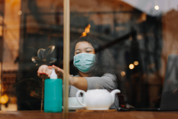 Attraktive junge Frau in medizinischer Maske mit antiseptischen Mitteln für die Hände, während sie im Stadtcafé sitzt. Konzept zum Schutz vor Viren während der Pandemiezeit. - Foto, Bild