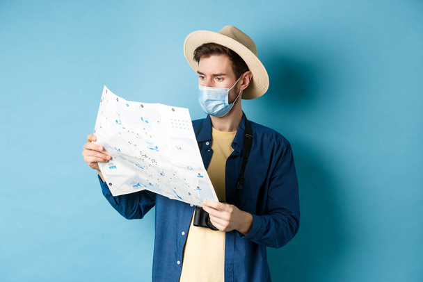Covid-19, pandemia i koncepcja podróży. Przystojny młody facet w masce i letniej czapce patrzący na mapę turystyczną, podróżujący na wakacje, stojący na niebieskim tle - Zdjęcie, obraz