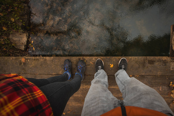 Две ноги в спортивной обуви стоят на деревянном мосту над спокойной водой с капельками дождя. Камера смотрит вниз к ногам и полу. - Фото, изображение