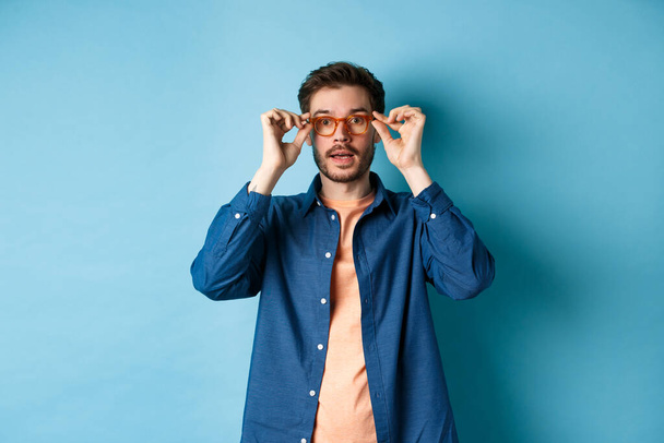 Bild des gutaussehenden kaukasischen Mannes setzt eine Brille auf und schaut in die Kamera, probiert neue Brillen im Optikergeschäft aus, blauer Hintergrund - Foto, Bild