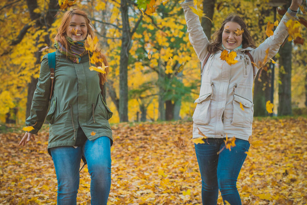 Две женщины бросают осенние листья в воздух в романтическом лесу. Две девушки играют с листьями в лесу. Романтическое осеннее фото. - Фото, изображение