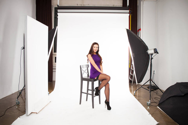 Portrait of a full length young beautiful brunette woman in a short purple dress sits on a chair, interior photo studio - Foto, Imagem