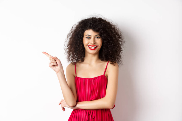 Hermosa mujer feliz con peinado rizado, con vestido festivo, señalando el dedo a la izquierda en el logotipo y sonriendo, fondo blanco - Foto, imagen
