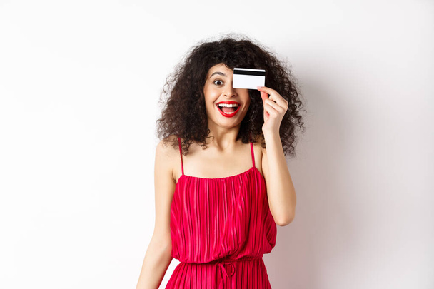 Alışverişe. Kırmızı elbiseli heyecanlı kıvırcık kız, plastik kredi kartı gösteriyor ve sevinçten haykırıyor, indirimlere ve fiyatlara hayran, beyaz arka planda duruyor. - Fotoğraf, Görsel