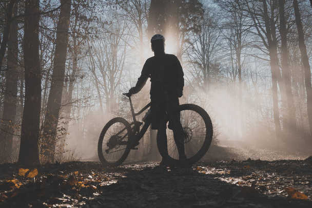 MTB o ciclista de montaña posando en un bosque brumoso con sol detrás de él. Silueta de un ciclista de montaña en los bosques de niebla, hermosa silueta deportiva - Foto, imagen