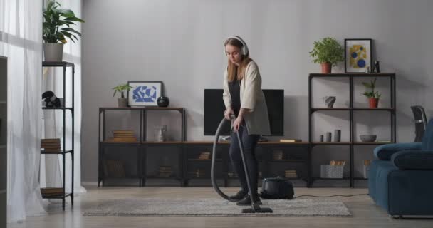 pretty teen girl is doing housework playfully and joyfully, hoovering carpet and dancing, listening to song in headphones - Footage, Video