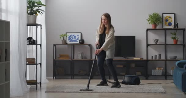teenager girl is cleaning home and dancing with vacuum cleaner in living room, fun and housework at weekend - Footage, Video