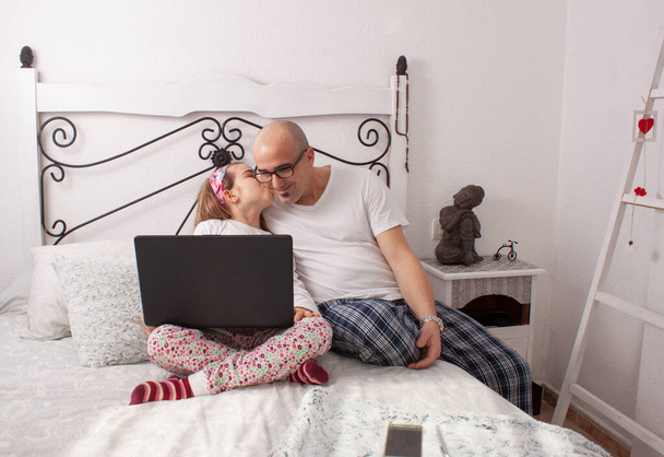 Homem em uma cama de casal com sua filha fazendo uma chamada de vídeo em um laptop uma manhã e ela beija seu pai. - Foto, Imagem
