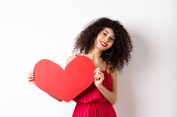 Mujer romántica en vestido mostrando gran corazón rojo, enamorándose, sonriendo feliz a la cámara, fondo blanco - Foto, Imagen