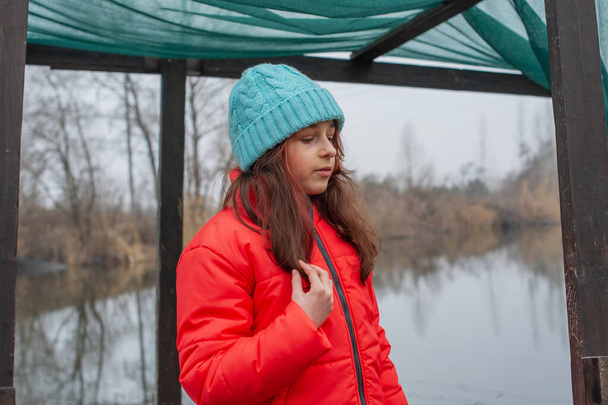 Chica en ropa de invierno sobre un fondo del río. Chica de 10 años. Adolescente en chaqueta naranja - Foto, imagen