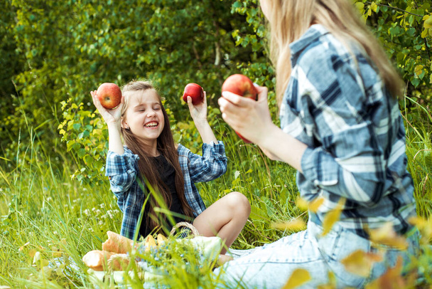 Mother with daughter in the park. mother holding the red apples near the eyes.summer park, fruit basket,white bread. outdoors. Family holidays and togetherness - 写真・画像