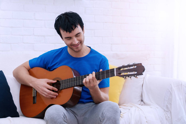 Привлекательный красивый молодой человек, сидящий в гостиной и играющий на акустической гитаре и поющий песни. Концепция образа жизни - Фото, изображение