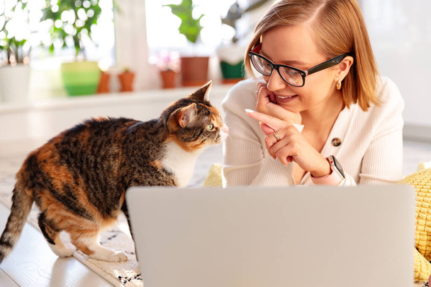 Mosolygó nő szabadúszó fekszik szőnyegen a nappaliban, dolgozik a laptop otthon során az önelkülönítés, macska a közelben akar figyelmet, és hogy stroke. Karantén, távoli munkavégzés. - Fotó, kép
