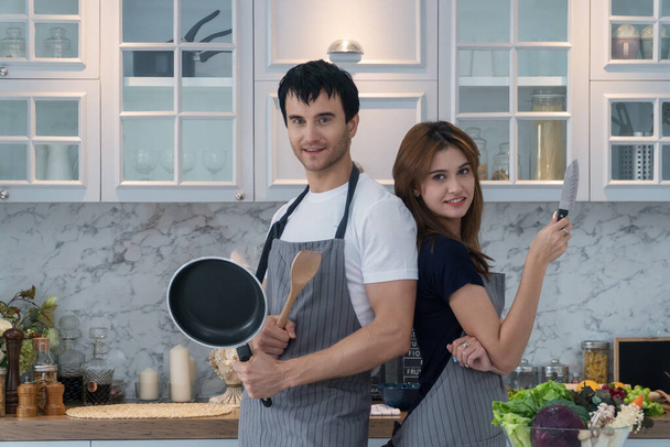 Lovely couple standing back to back in kitchen. Smiling handsome young man wear an apron while holding frying pan and wooden spoon his girlfriends holding knife with vegetables looking at camera.  - Photo, Image