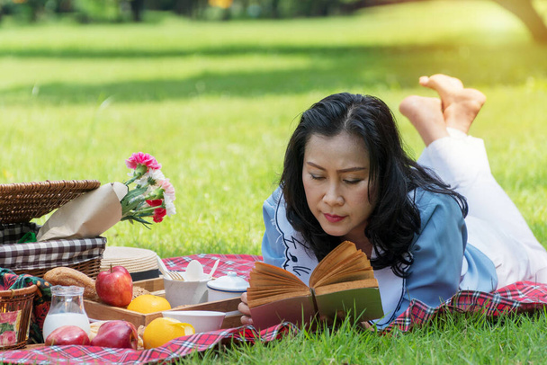 高齢者のライフスタイルコンセプト。シニア女性50読書魅力的な本で彼女の手にピクニックと余暇時間を持っている公共公園で緑の芝生の上でリンゴ、オレンジ、ミルクとパン . - 写真・画像