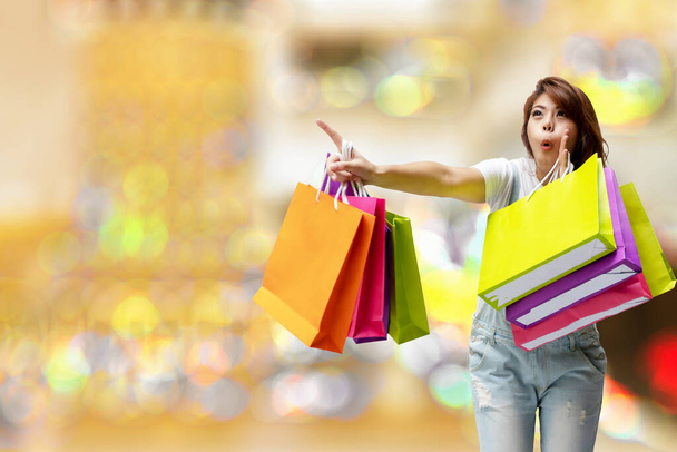 Vidám izgatott fiatal nő kezében színes bevásárló táskák ujjal mutogató bokeh könnyű puha hatású háttér. Boldogság, fogyasztás, értékesítés és az emberek koncepciója. - Fotó, kép