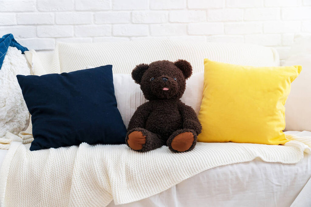 Medvídek tmavě hnědý na bílém gauči s modrým polštářem a žlutým polštářem. Medvídek hravý na bílé pohovce v obývacím pokoji. - Fotografie, Obrázek