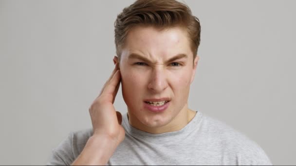 Ωτίτιδα. Πορτρέτο του νεαρού άνδρα που υποφέρει από πόνο στο αυτί, κοντινό πλάνο, γκρι φόντο - Πλάνα, βίντεο