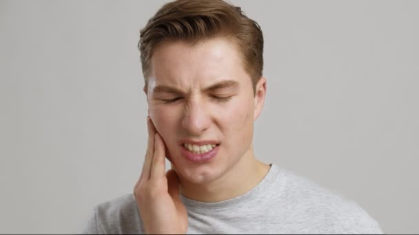 Tandpijn. Jongeman die lijdt aan acute kiespijn, wrijven zijn pijnlijke wang, grijze studio achtergrond - Video