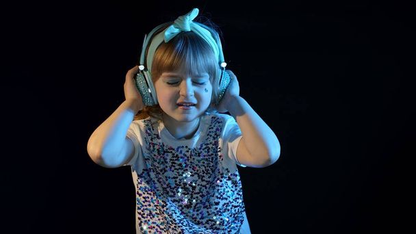 Stylish teenager kid girl standing among neon lights listening to music in headphones, dancing - Photo, Image