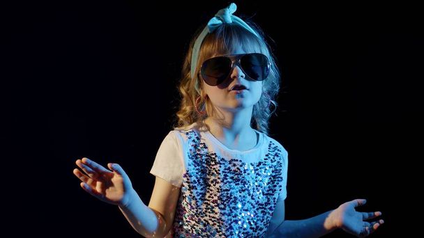 Chica de moda en gafas futuristas bailando, escuchando música en auriculares sobre fondo negro - Foto, Imagen