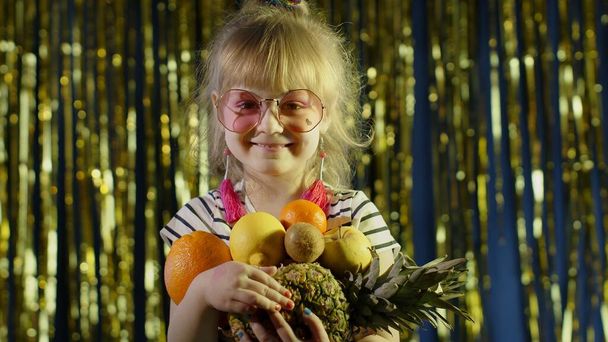 Trendy girl in pink sunglasses posing looking at camera with bunch of fruits in hands in night club - Zdjęcie, obraz