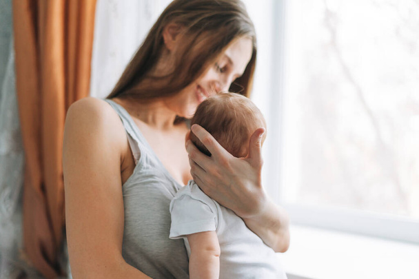 Mladá žena maminka s dlouhými vlasy drží dítě dívka na rukou v blízkosti okna doma - Fotografie, Obrázek