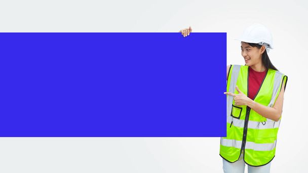 An Asian female engineer wearing a green vest cloth and white helmet stood holding a blue paper placard with empty space for text, on isolated background. - Foto, imagen